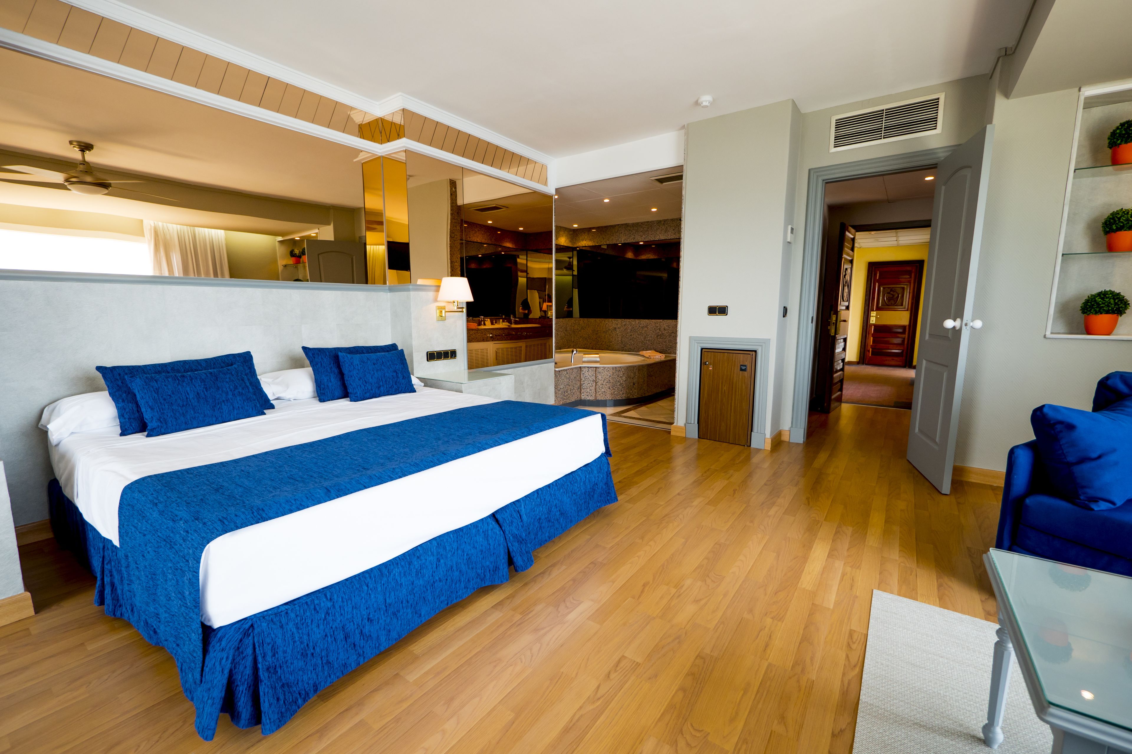 Una habitación de hotel moderna con una cama grande, una zona de estar con un sofá azul, una mesita y un baño con bañera y ducha.