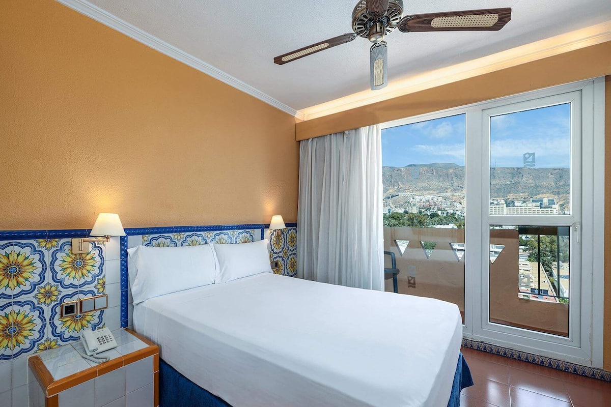 Ein Hotelzimmer mit einem Bett, einem Nachttisch mit Telefon, einem Deckenventilator und Blick auf eine Stadtlandschaft durch große Fenster.