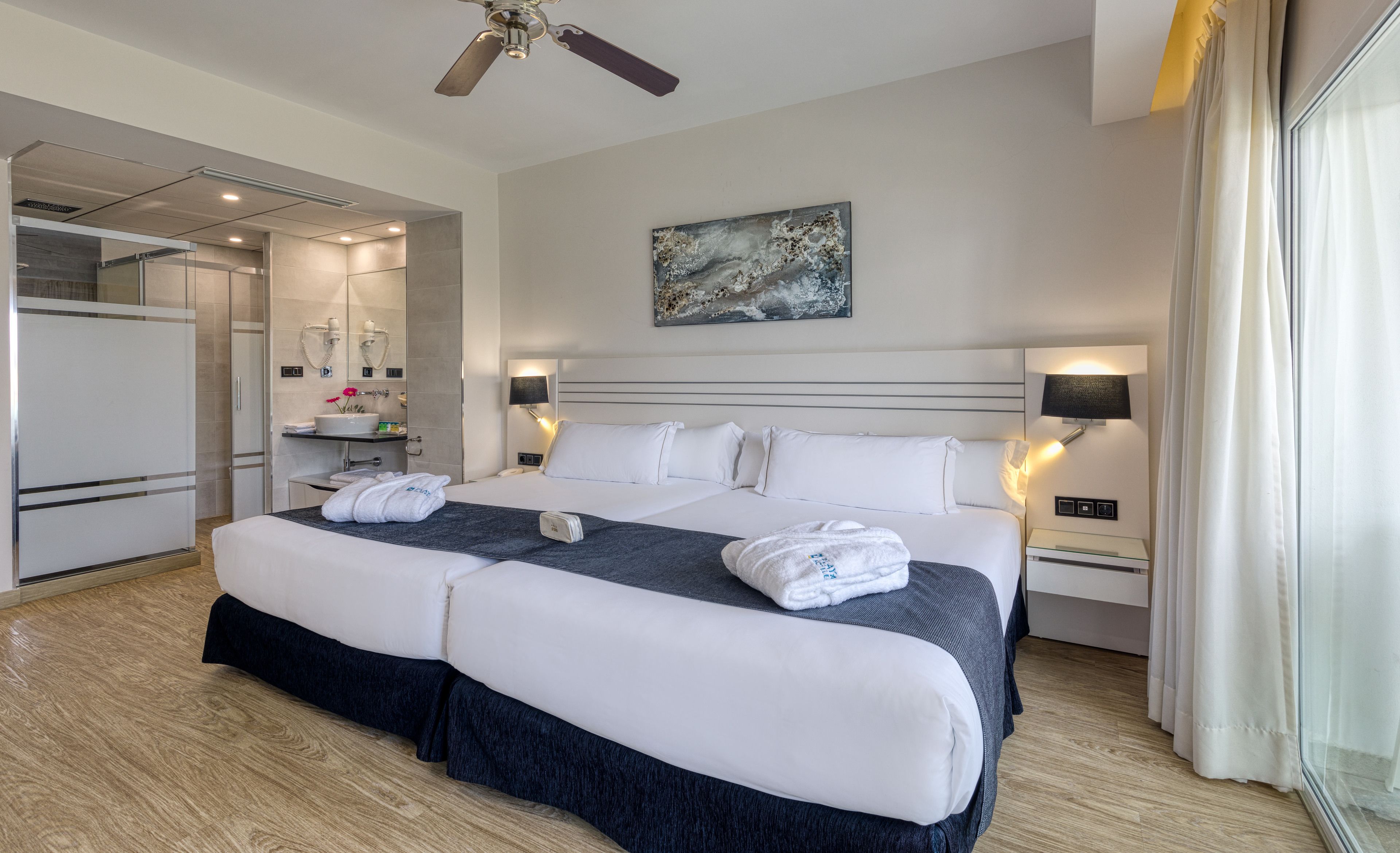 A modern hotel room with a large bed, bedside lamps, a painting on the wall, and a bathroom visible through an open door.