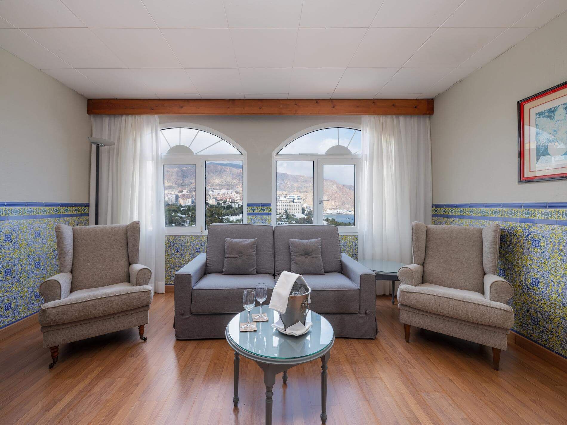 A room with a sofa, two armchairs, a glass table with a bottle and glasses, and large windows with curtains.