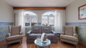 Habitación con sofá, dos sillones, una mesa de vidrio con botella y vasos, y ventanas grandes con cortinas.