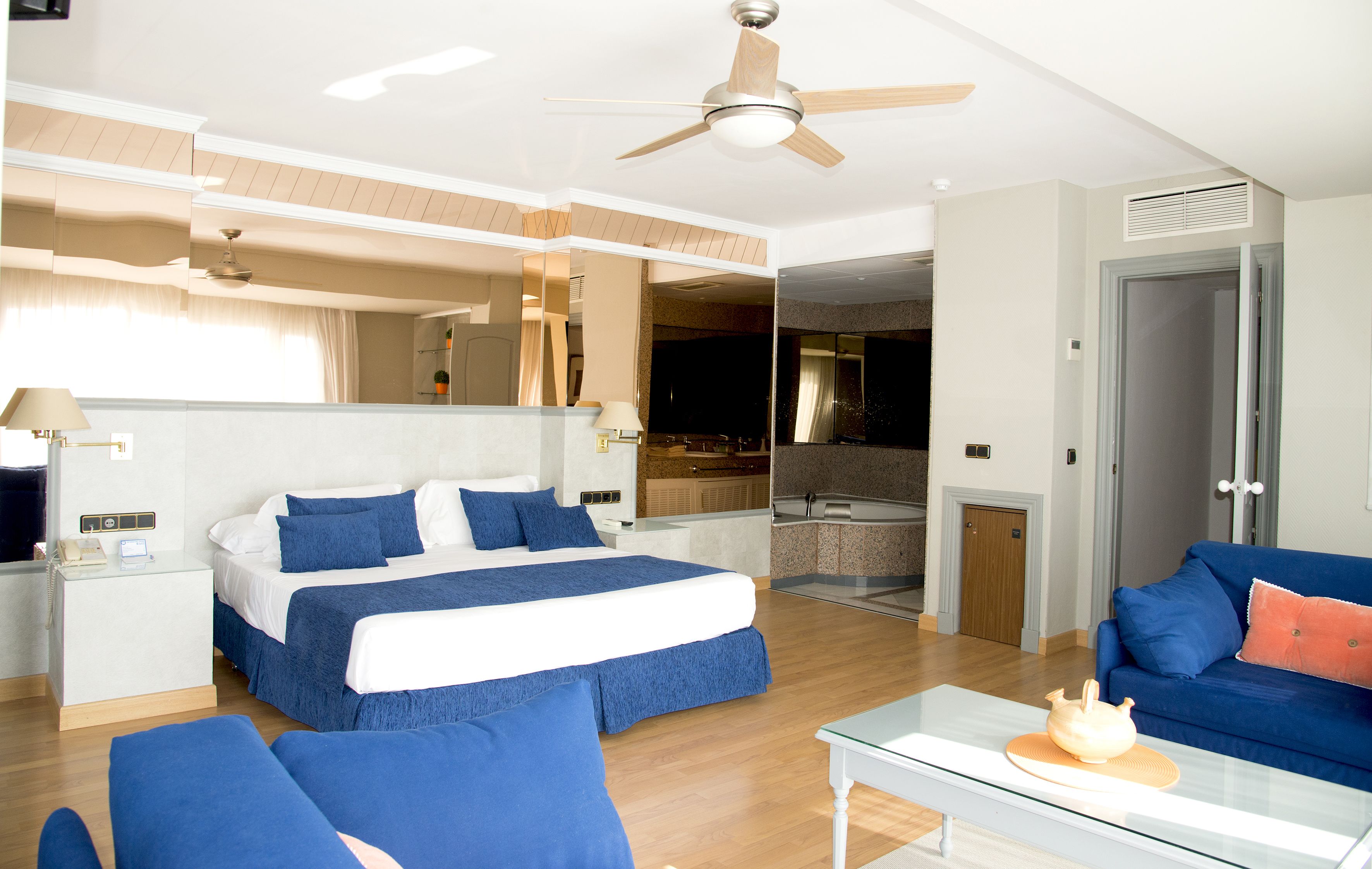 A hotel room with a bed, a sofa, a coffee table, a ceiling fan, and a bathroom visible through an open door.
