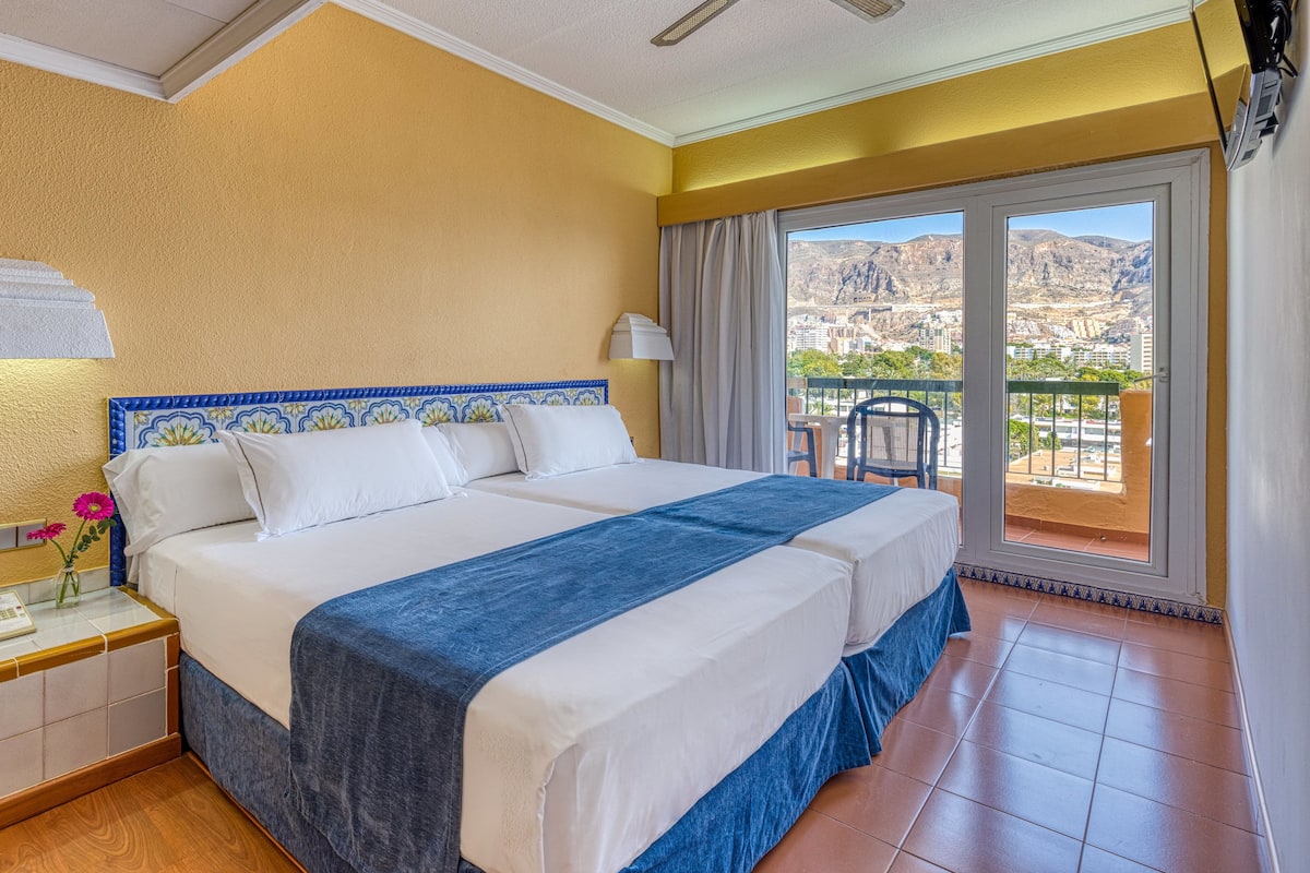 Ein Hotelzimmer mit einem großen Bett, einem Nachttisch mit Telefon und Blick auf die Berge vom Balkon aus.