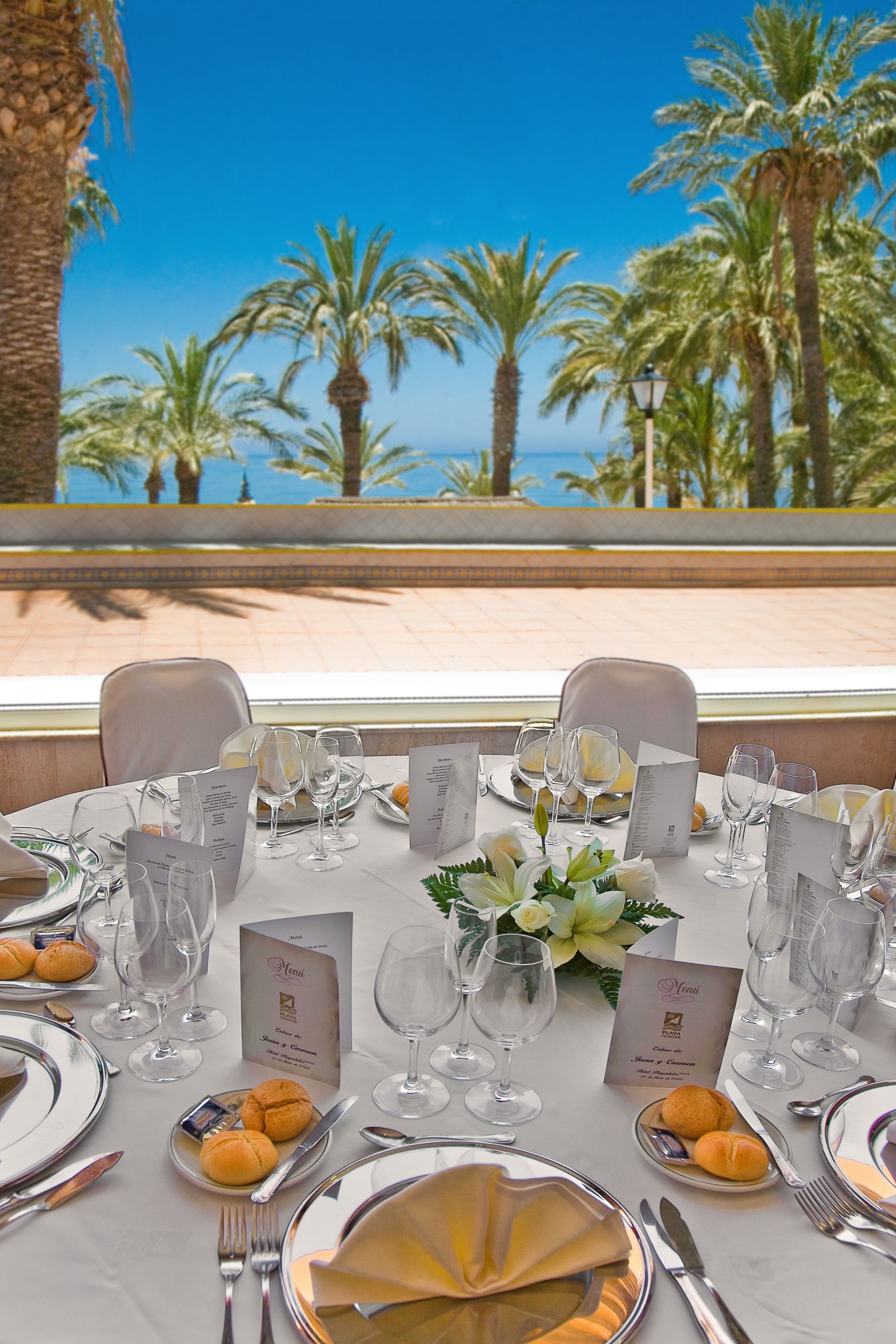 Een gedekte tafel met menu, glazen en borden, met uitzicht op een strand met palmbomen.