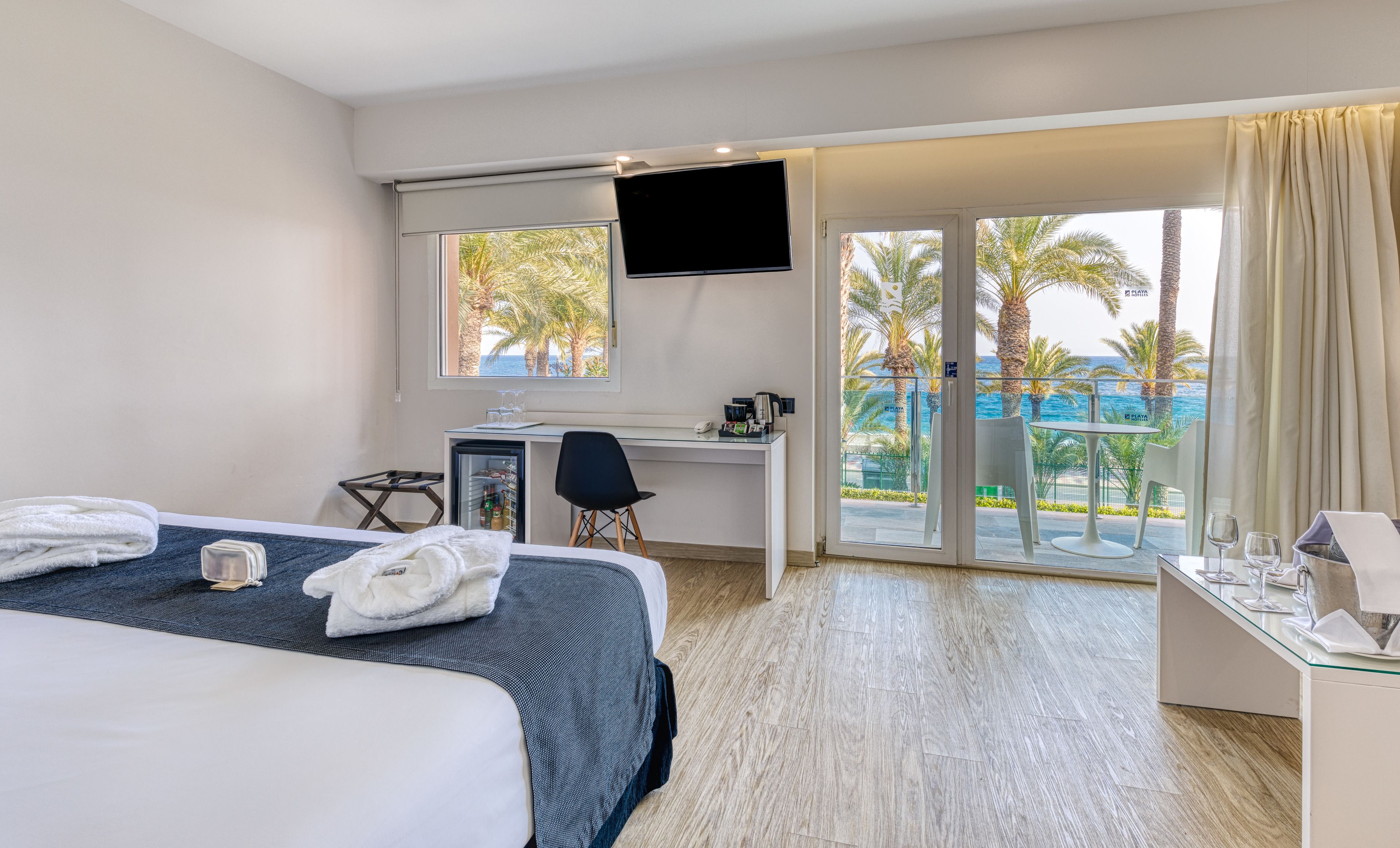 Habitación de hotel con cama, escritorio y balcón con vista a palmeras y al mar.