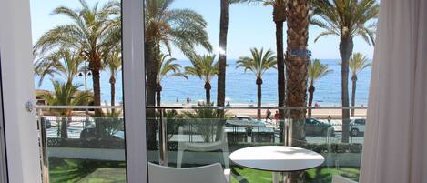 A balcony with a glass railing overlooking a beach with palm trees and the ocean. There are white chairs and a small table.