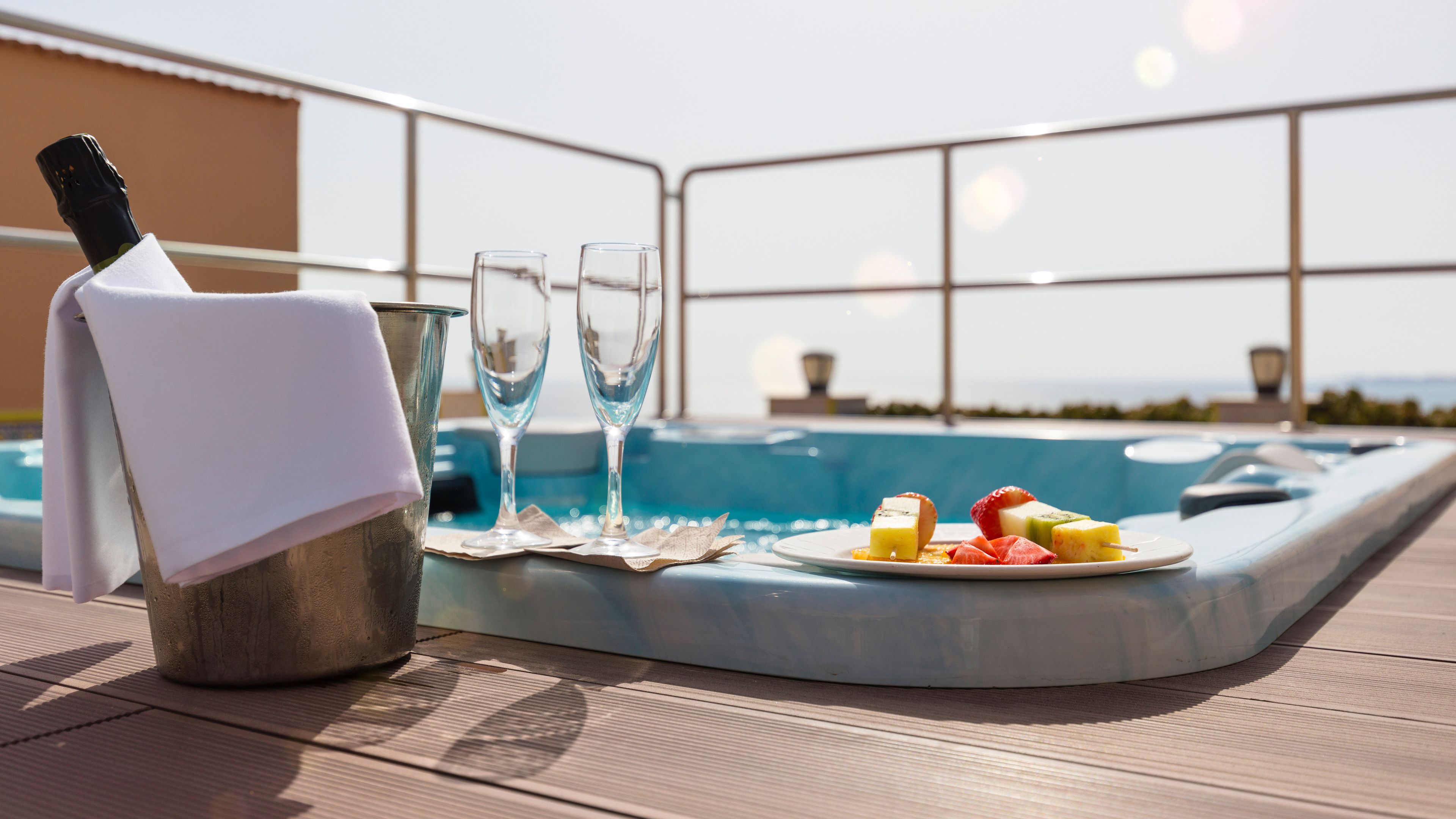Escena junto a la piscina con un jacuzzi, una botella de champán, dos copas y un plato de brochetas de fruta.