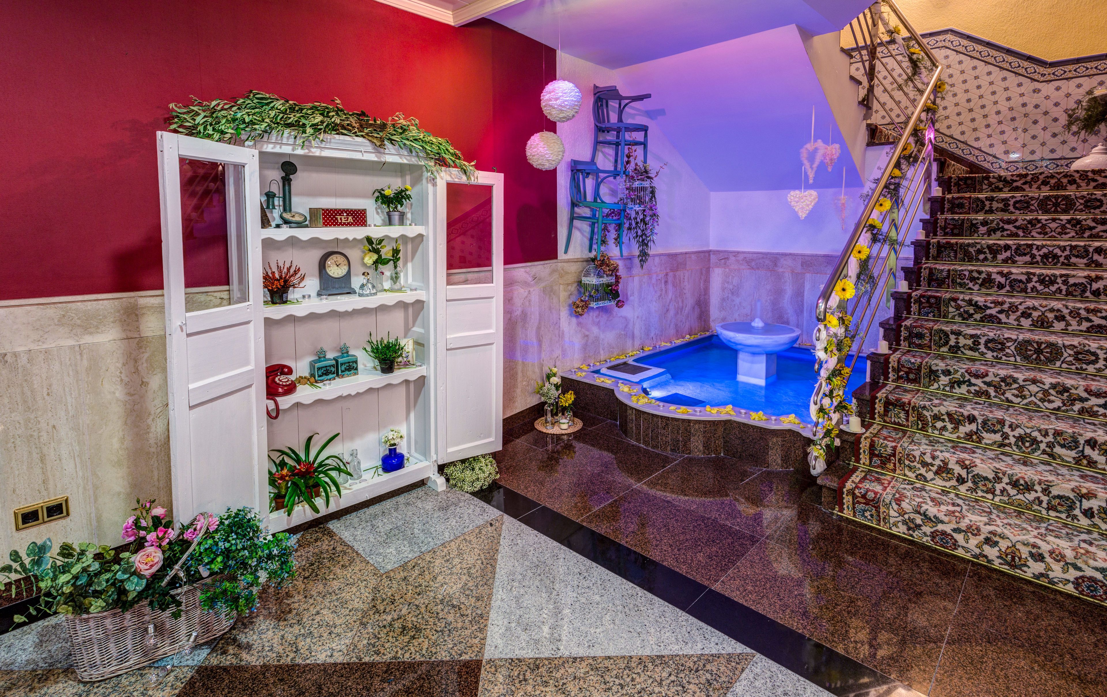 Un baño con bañera, un estante blanco con diversos objetos, una pared roja y una escalera con alfombra floral.