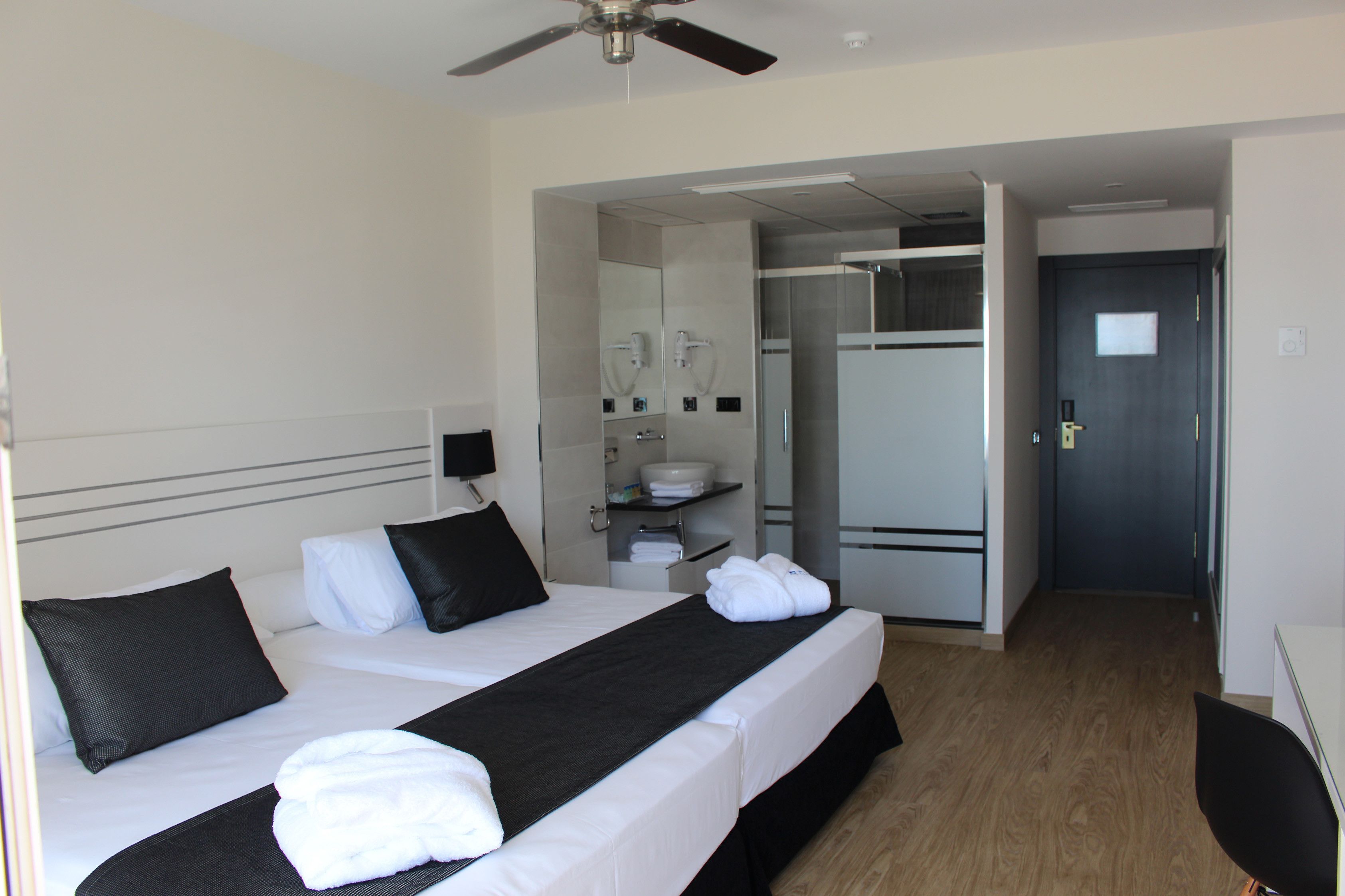 Hotelkamer met een bed, een bureau, een stoel, een plafondventilator, een badkamer met een wastafel en spiegel, en een deur.