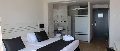 Habitación de hotel con cama, escritorio, silla, ventilador de techo, baño con lavamanos y espejo, y una puerta.