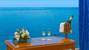 Una mesa con una botella de champán, dos copas y un arreglo floral frente a una ventana con vista al mar.