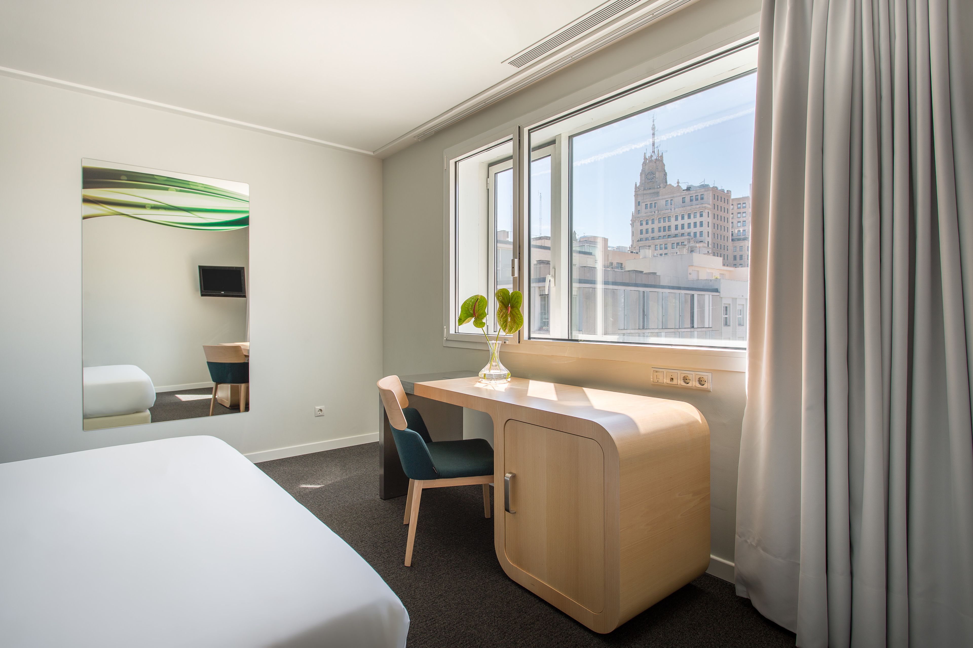 Una habitación de hotel con cama, escritorio, silla, una mesita, espejo y ventana con cortinas, y vista a la ciudad.