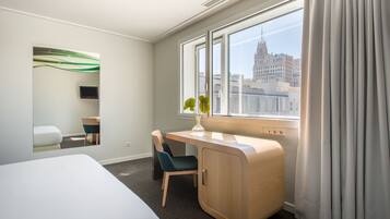 Una habitación de hotel con cama, escritorio, silla, una mesita, espejo y ventana con cortinas, y vista a la ciudad.