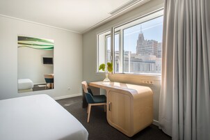 Ein Hotelzimmer mit Bett, Schreibtisch, Stuhl, kleinem Tisch, Spiegel und einem Fenster mit Vorhängen, mit Blick auf die Stadt.