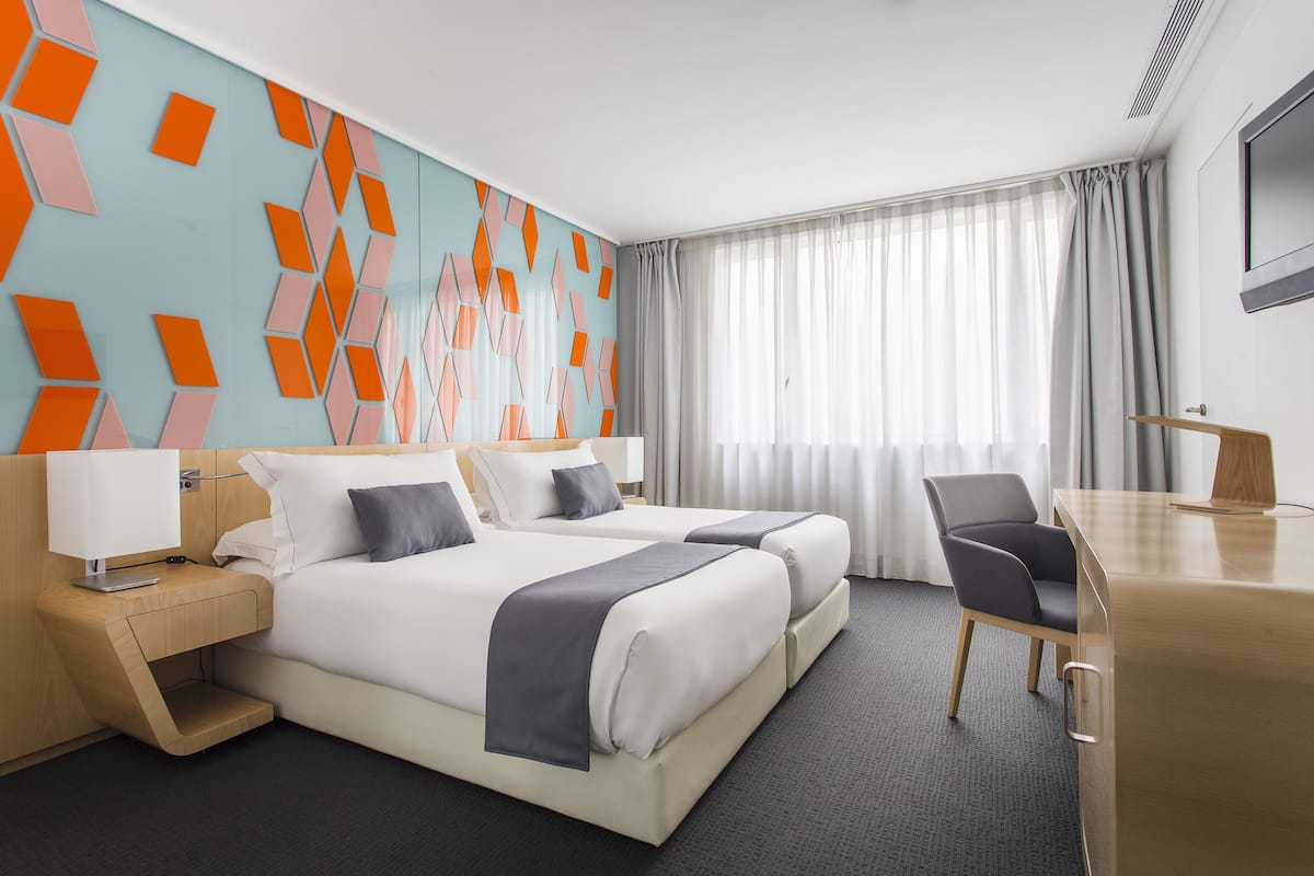 A hotel room with two beds, a desk, a chair, a TV, and a window with curtains.