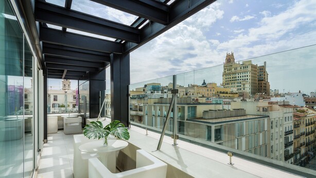Un balcón moderno con barandilla de vidrio, una planta en maceta y vistas a un paisaje urbano con edificios históricos.