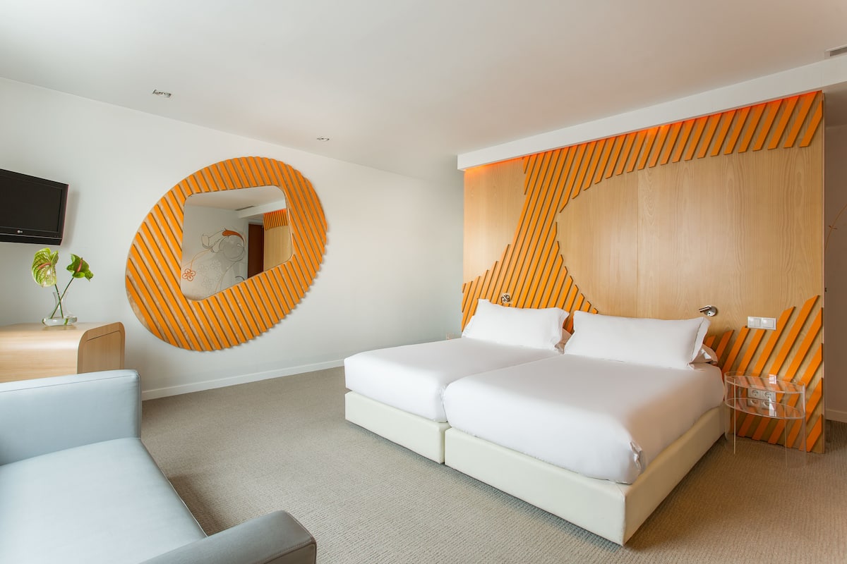 A modern hotel room with a large bed, a round mirror, a wooden headboard, and a small table with a plant.