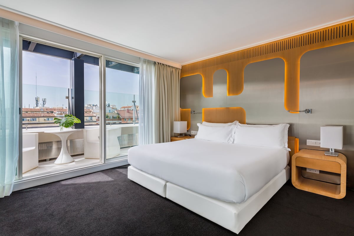 A modern hotel room with a large bed, a small table, a nightstand, and a view of the city through large windows.