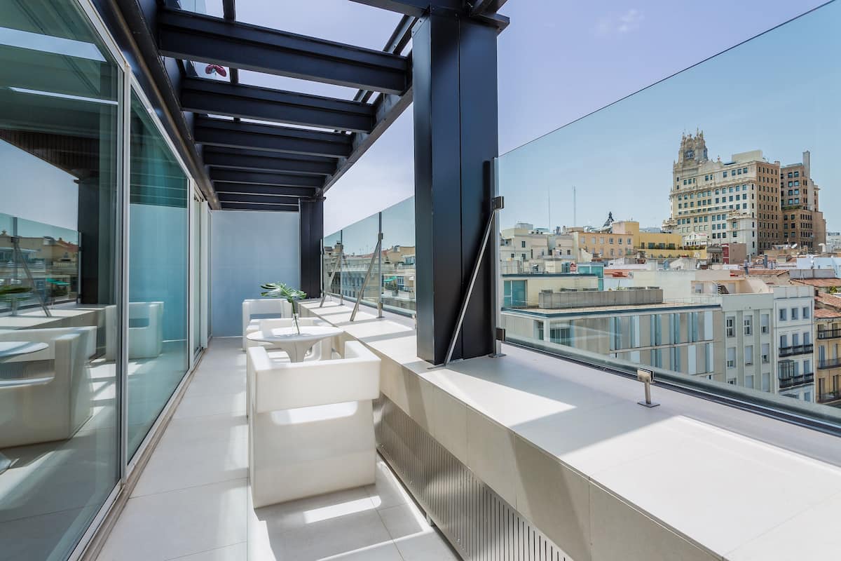 A modern rooftop terrace with glass railings, white furniture, and a city view.