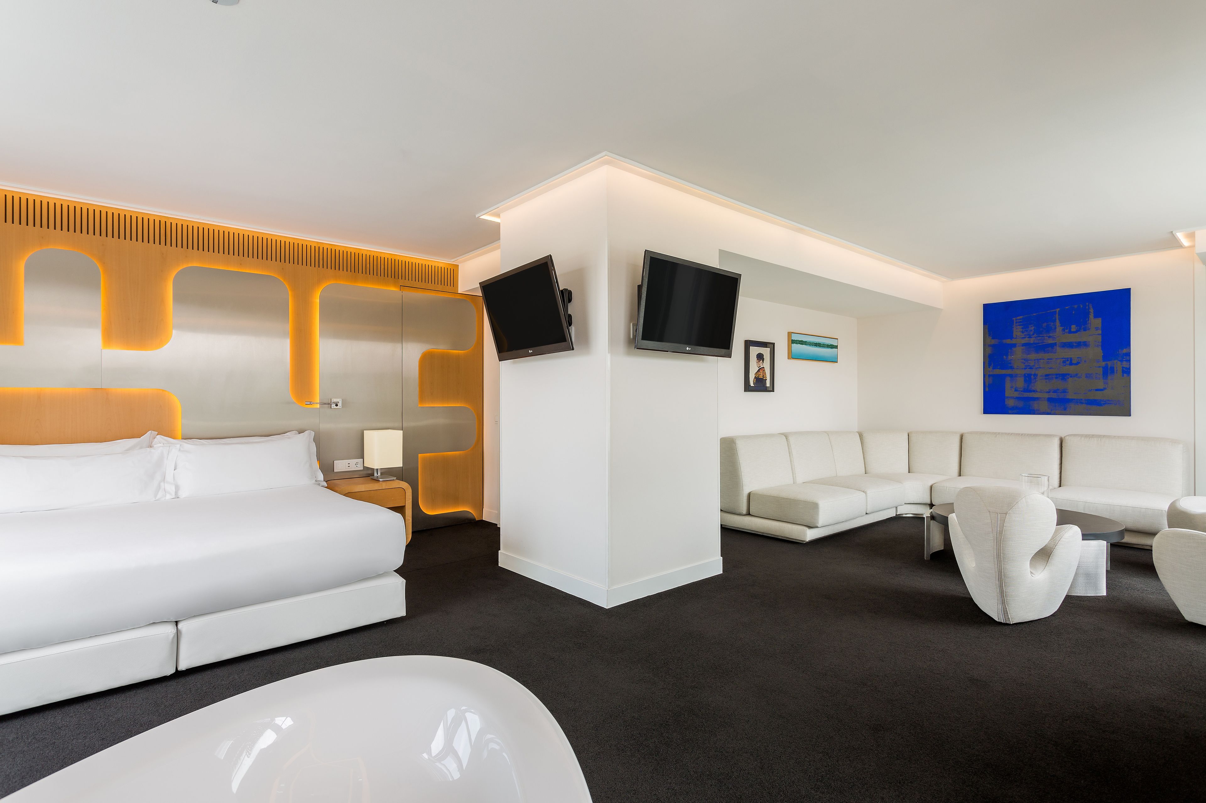 Una habitación de hotel moderna con una cama blanca, un sofá, una mesa de centro y un televisor fijado a la pared.