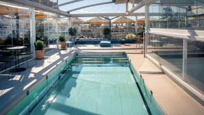 Una piscina en la azotea con paredes de vidrio, asientos exteriores y vistas a la ciudad.