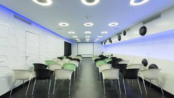 Une salle de conférence avec des rangées de chaises disposées face à un écran de projection, équipée de luminaires au plafond et d’un système de ventilation.