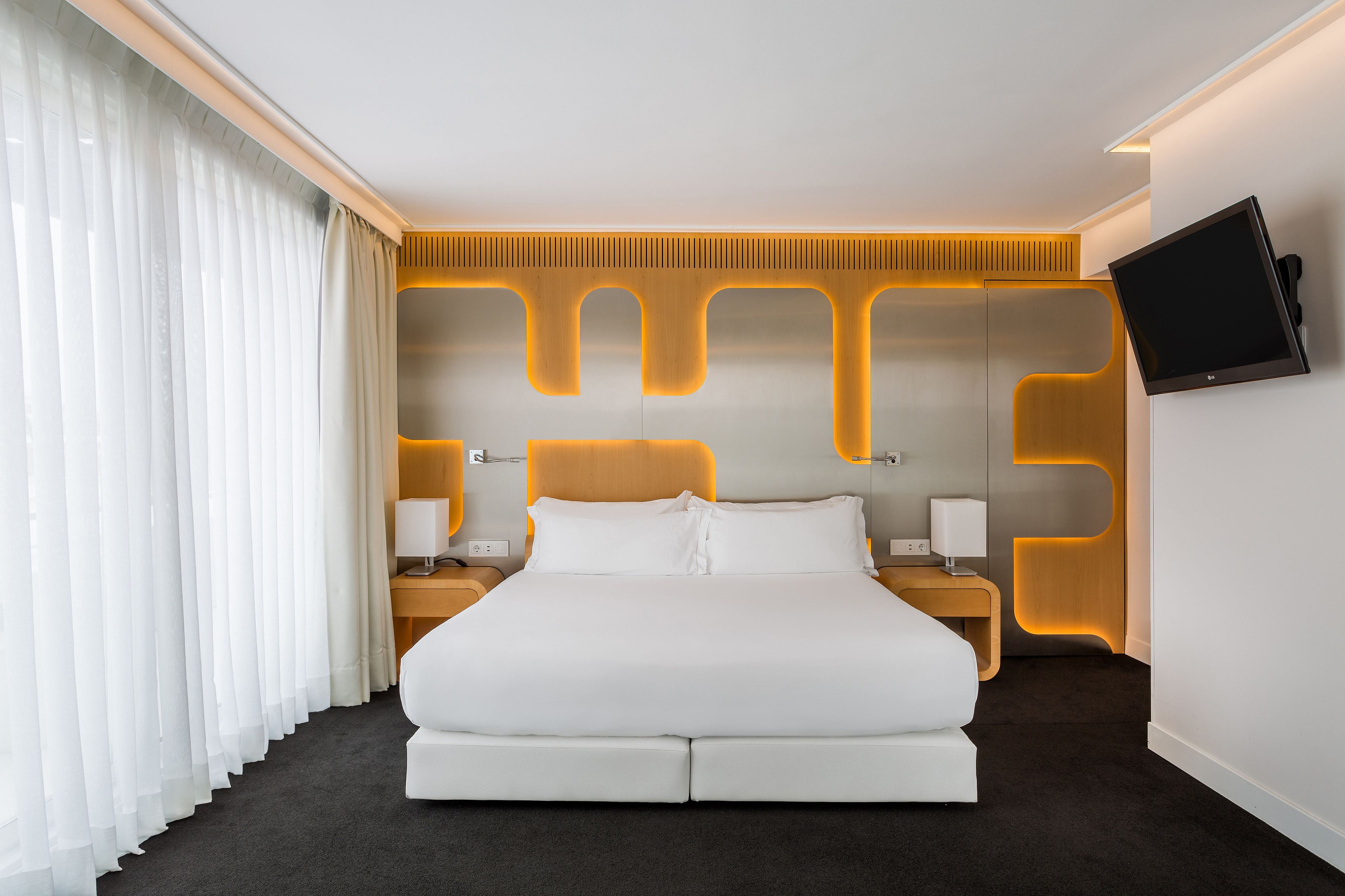 A modern hotel room with a large bed, a flat-screen TV mounted on the wall, and a wooden accent wall.