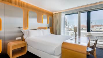 Habitación de hotel moderna con una cama grande, una mesita de noche de madera, un escritorio con una silla y un ventanal con vistas a la ciudad.