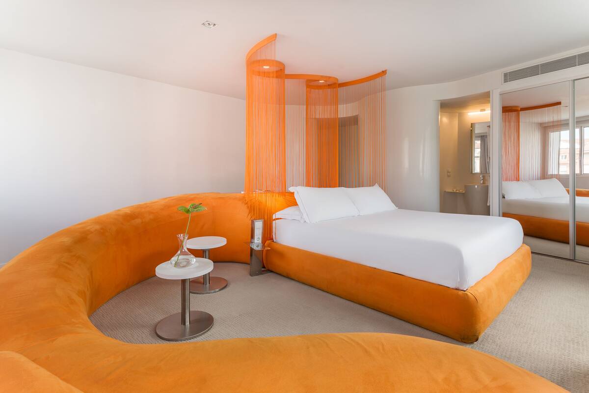 A modern hotel room with a curved orange sofa, a small round table, a bed with white linens, and a large mirror.