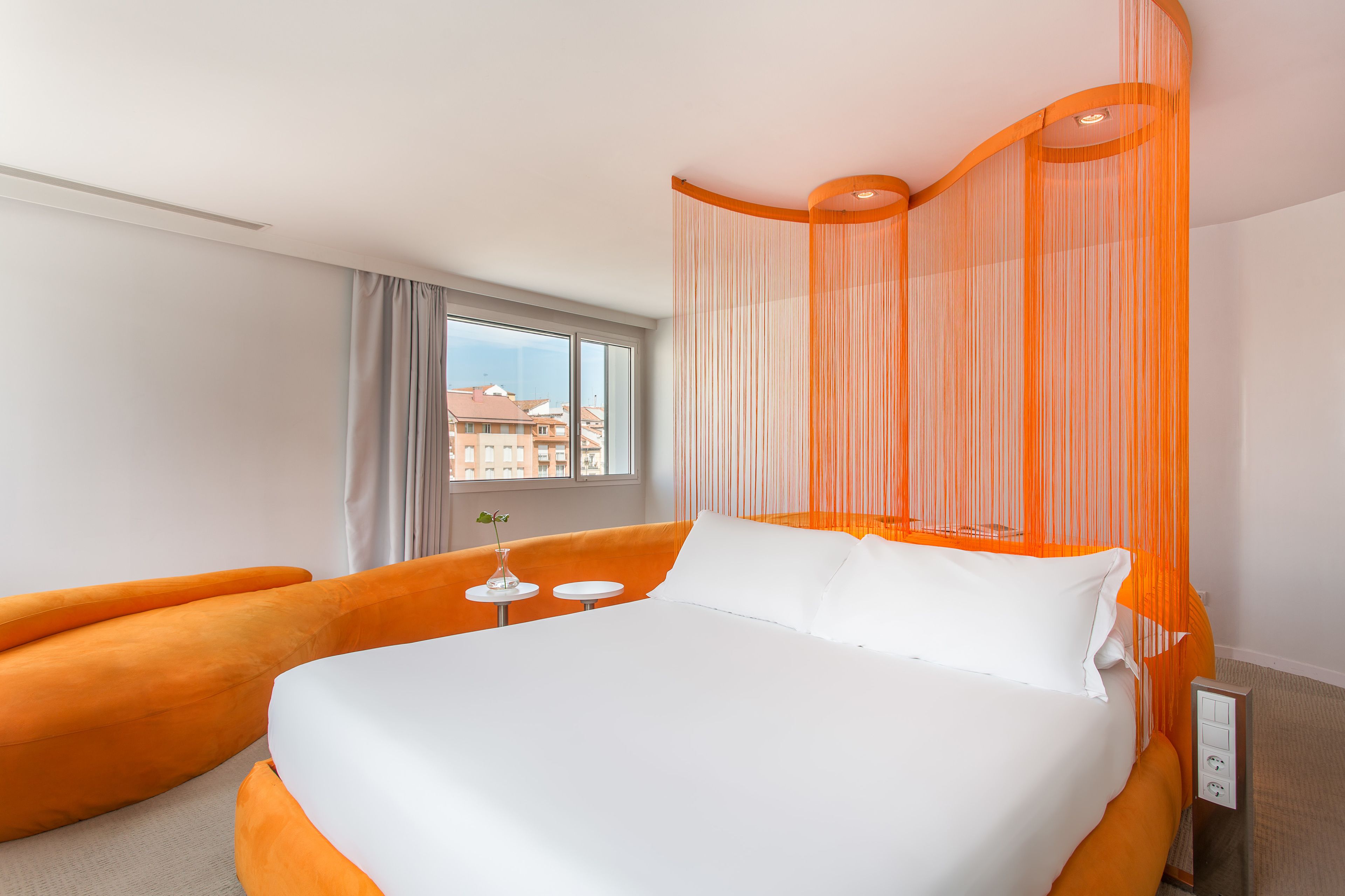 Habitación de hotel con una cama grande, almohadones color naranja, una mesita con un florero y una ventana con vistas a edificios.