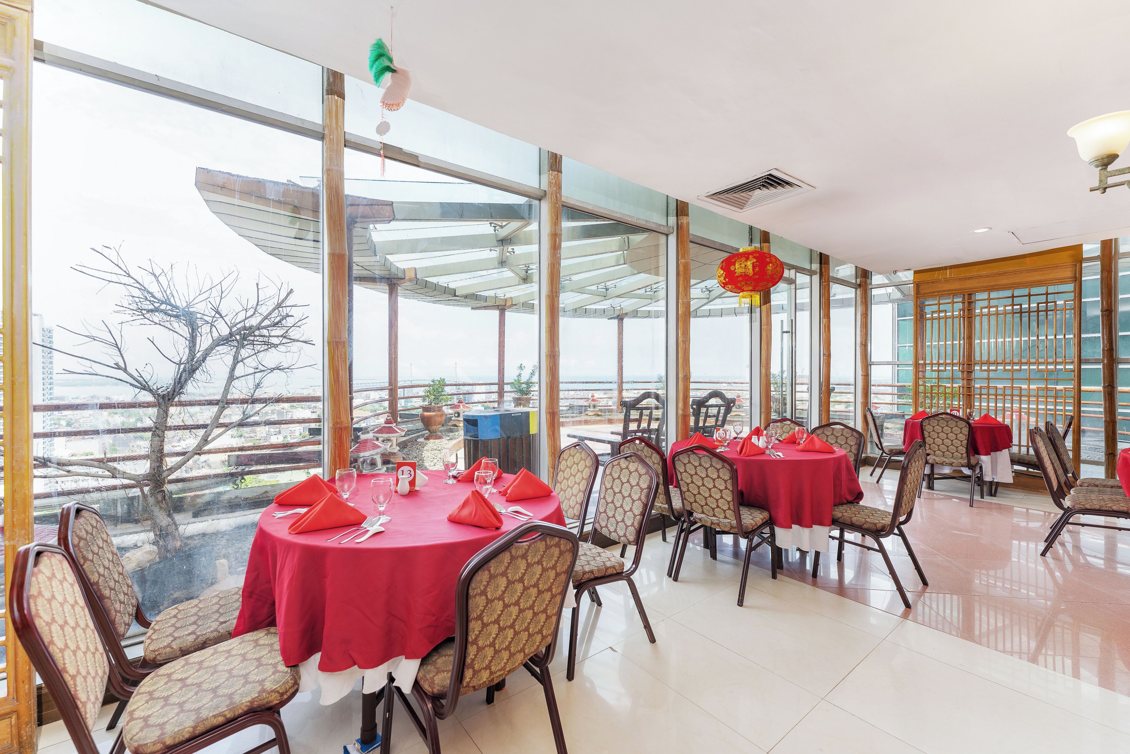 A restaurant with round tables covered in red tablecloths, wooden chairs, and large windows offering a view of the outside.