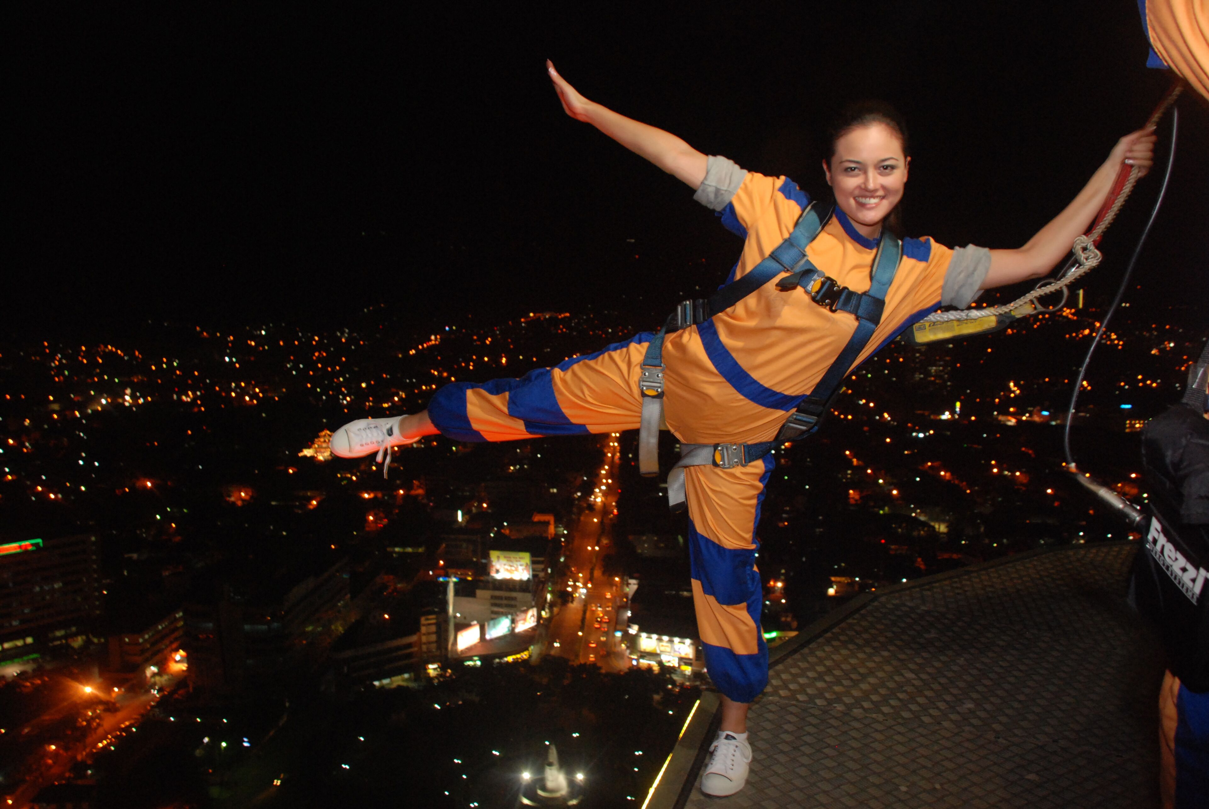 A person bungee jumping with a harness, wearing a yellow and blue outfit, with a cityscape in the background.