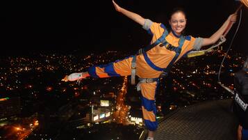 A person bungee jumping with a harness, wearing a yellow and blue outfit, with a cityscape in the background.