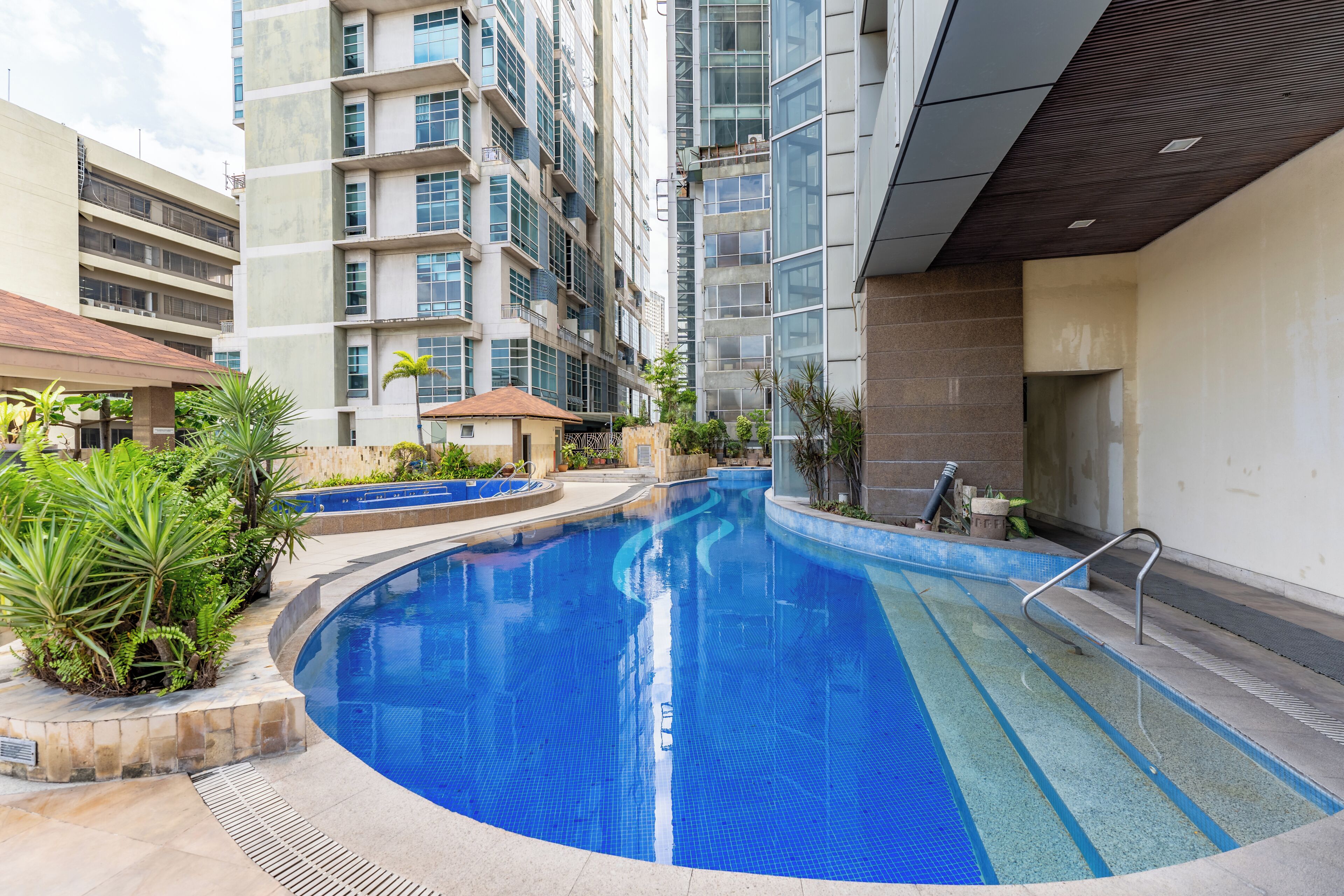 Una piscina rodeada de un área ajardinada con plantas y un edificio de varios pisos al fondo.