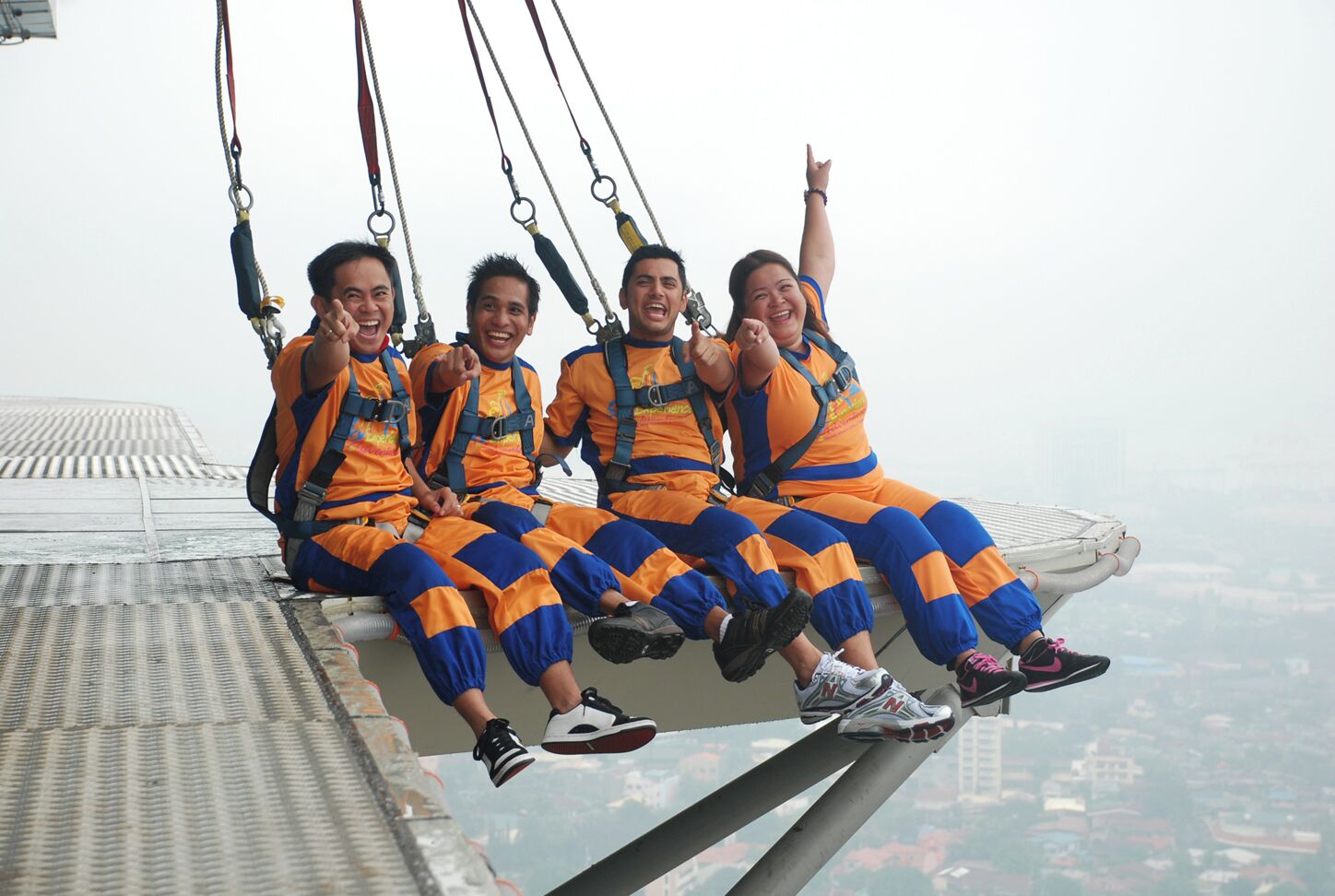 Four people sitting on a high platform with safety harnesses, overlooking a cityscape.