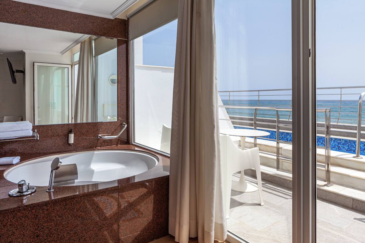 Une salle de bain moderne avec un grand lavabo et un miroir, un balcon avec une piscine et un fauteuil, et une vue dégagée sur l’océan.