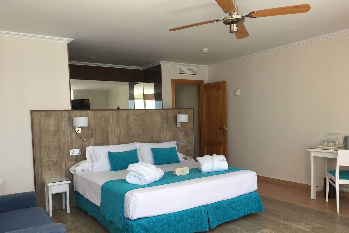 Une chambre d’hôtel avec un lit, un ventilateur de plafond, une table de chevet et un petit bureau avec une chaise.