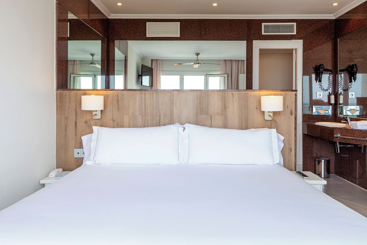 Une chambre d’hôtel avec un grand lit, deux lampes de chevet, une tête de lit en bois, un miroir, un ventilateur de plafond et une salle de bain avec un lavabo et un porte-serviettes.