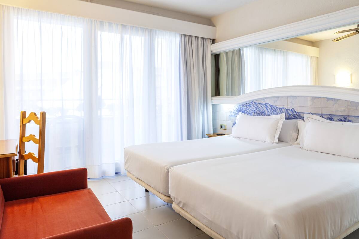 Une chambre d’hôtel avec deux lits, une chaise en bois, une petite table et une grande fenêtre avec des rideaux.