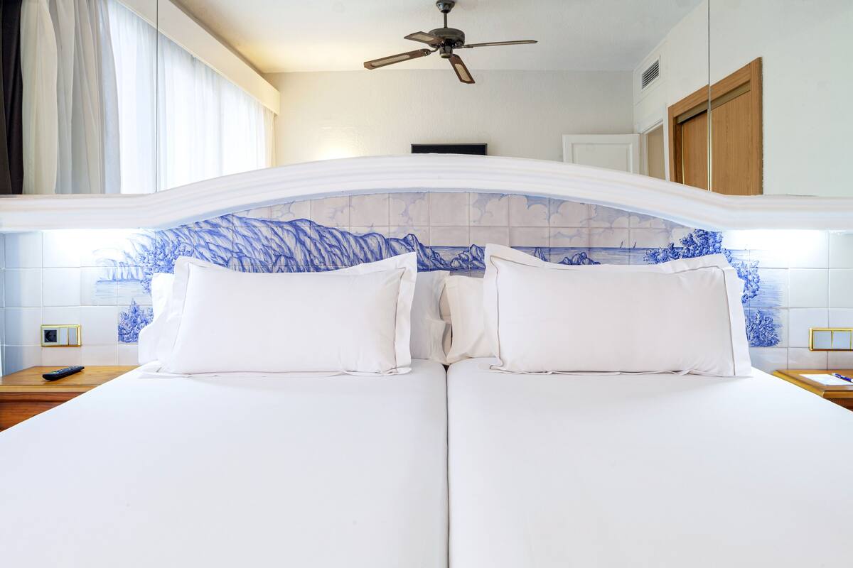 Un lit double avec du linge de lit blanc, un ventilateur de plafond et une tête de lit décorative en carreaux bleus.
