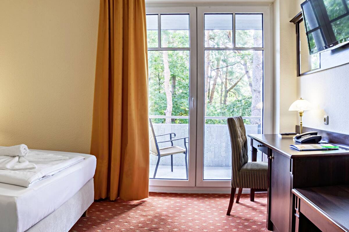 Una habitación de hotel con una cama, dos lámparas de noche, un escritorio con una silla y un ventanal con cortinas.