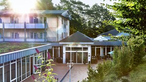 A reception area with a glass door entrance, a paved courtyard, and a building with multiple floors and balconies. - Ostseehotel Villen Im Park – ein Rovell Hotel (Heringsdorf)