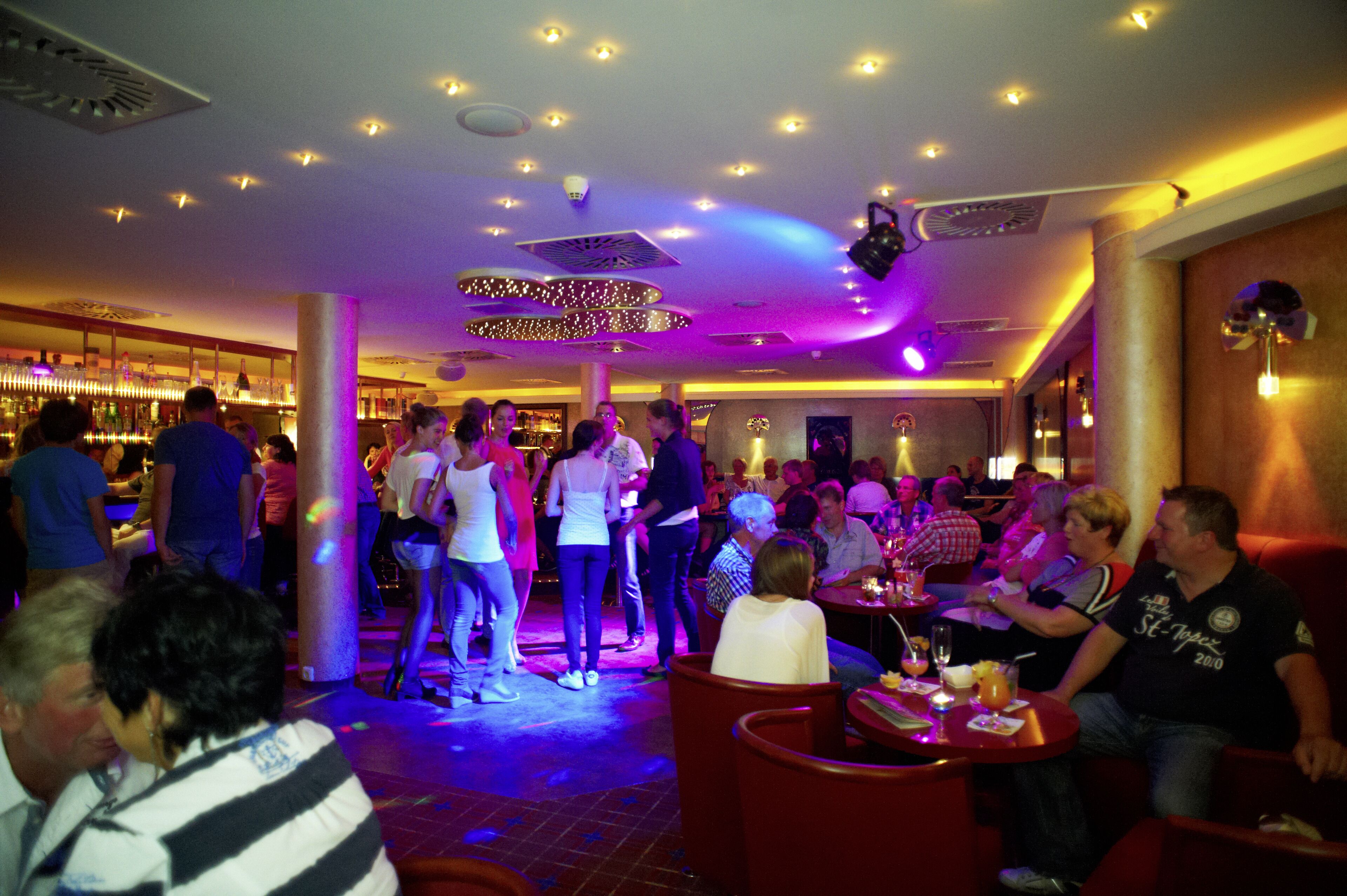 A crowded indoor venue with people standing and sitting, illuminated by ceiling lights and decorative lighting.