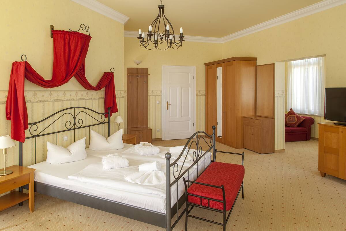 Una cama bien hecha con sábanas blancas y cortinas rojas, un armario de madera, un banco rojo y un televisor sobre un mueble de madera.