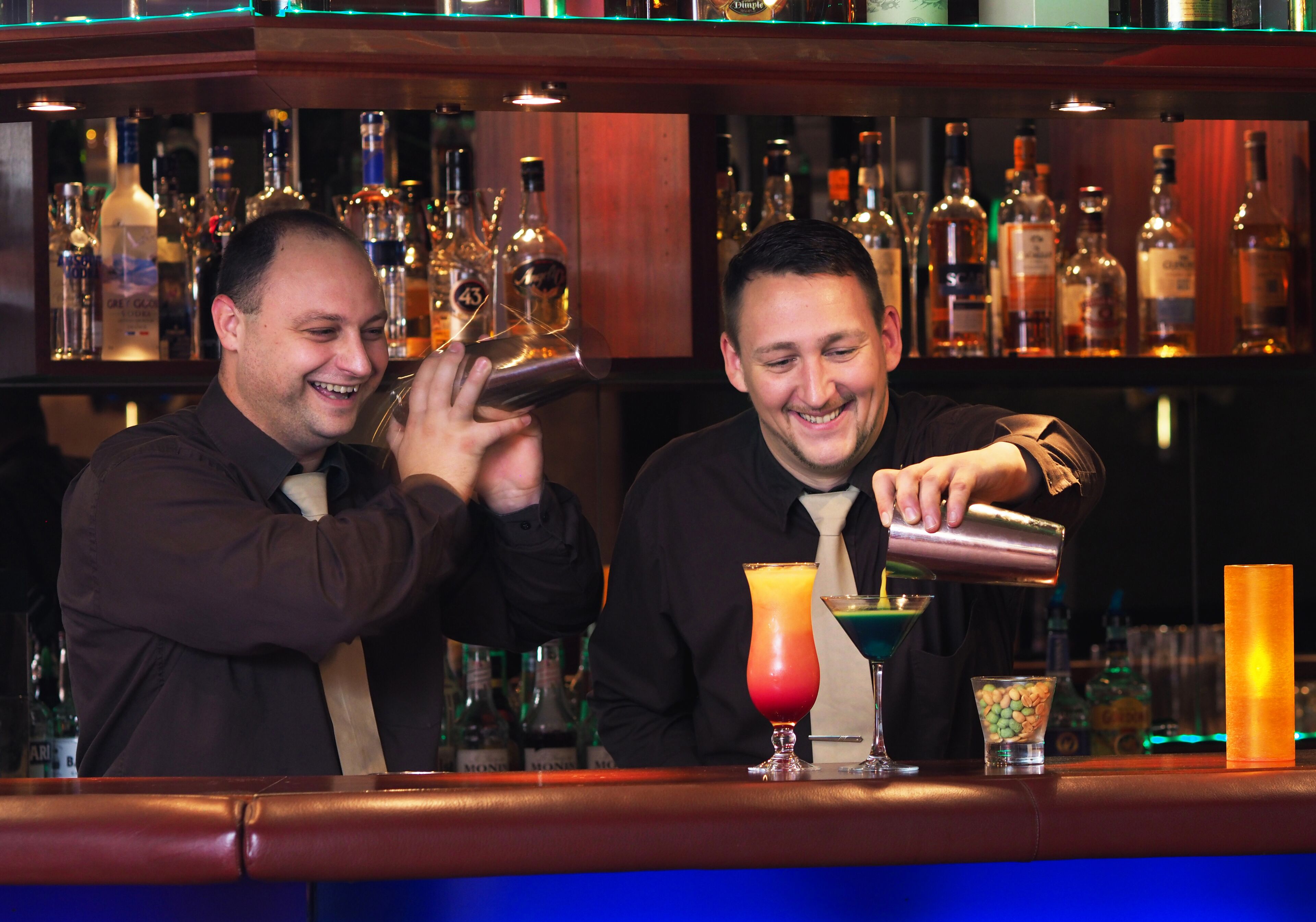 Dois bartenders preparando bebidas atrás do balcão, com diversas garrafas e copos.