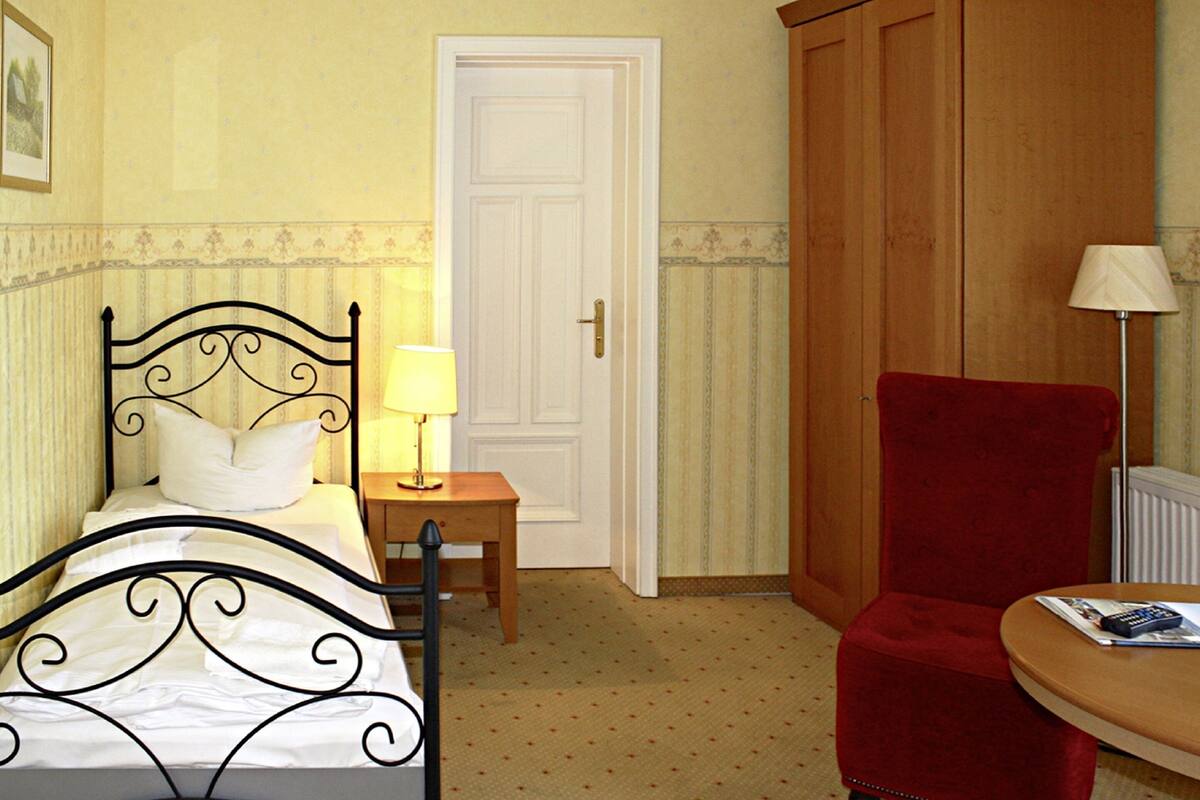 Una habitación con una cama, una mesita de noche con una lámpara, una silla roja y un armario de madera.
