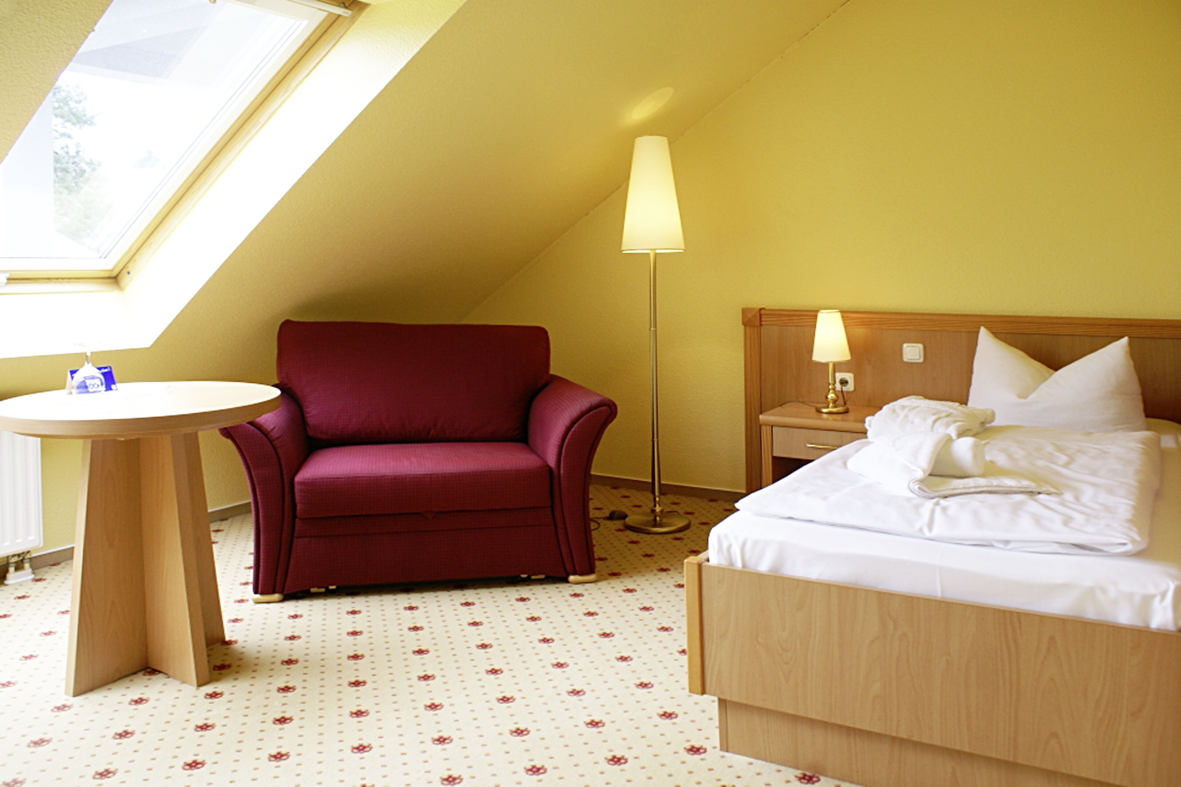 Ein Hotelzimmer mit einem Bett, einem roten Sessel, einem runden Tisch und einer Stehlampe.