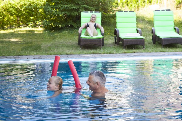 Una piscina con dos personas nadando y sillas de descanso verdes al fondo.