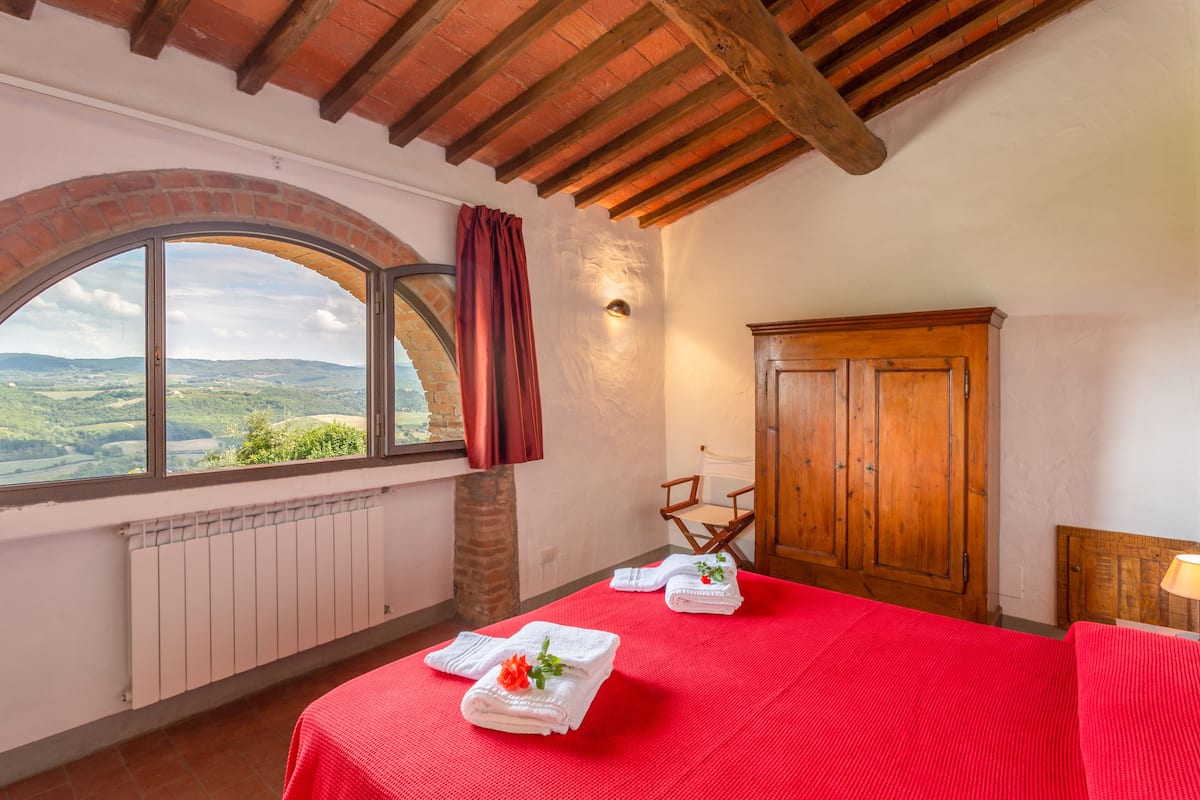 A room with a red bedspread, a wooden wardrobe, a window with a view of the countryside, and a radiator.