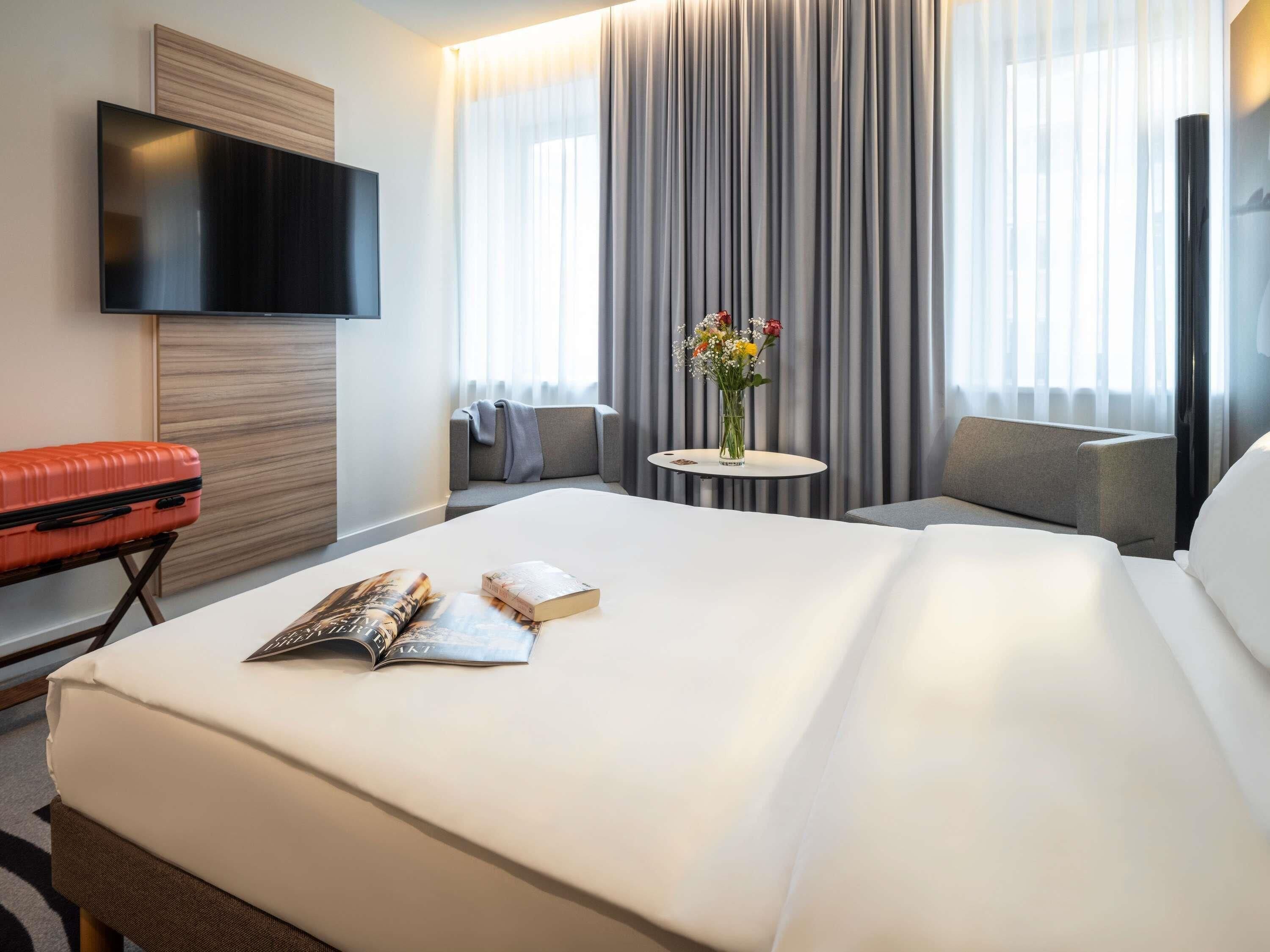 A hotel room with a large bed, a flat-screen TV mounted on the wall, a small round table with a vase of flowers, and a grey sofa.